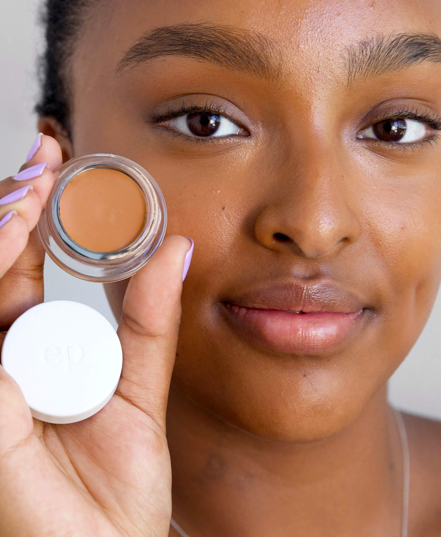 retrato de mujer viendo a la camara sonriendo con la piel maquillada sosteniendo la base corrector de arnica en tono mocha ere perez disponible en blumma