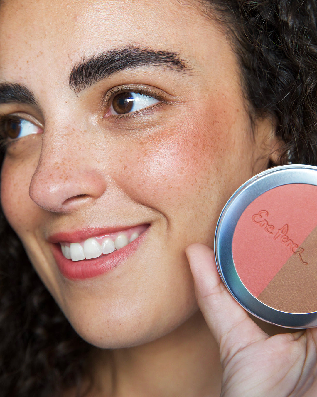 mujer sonriendo con tono brooklyn en sus mejillas de blush y bronceador ere perez  sosteniendo envase disponible en Blumma
