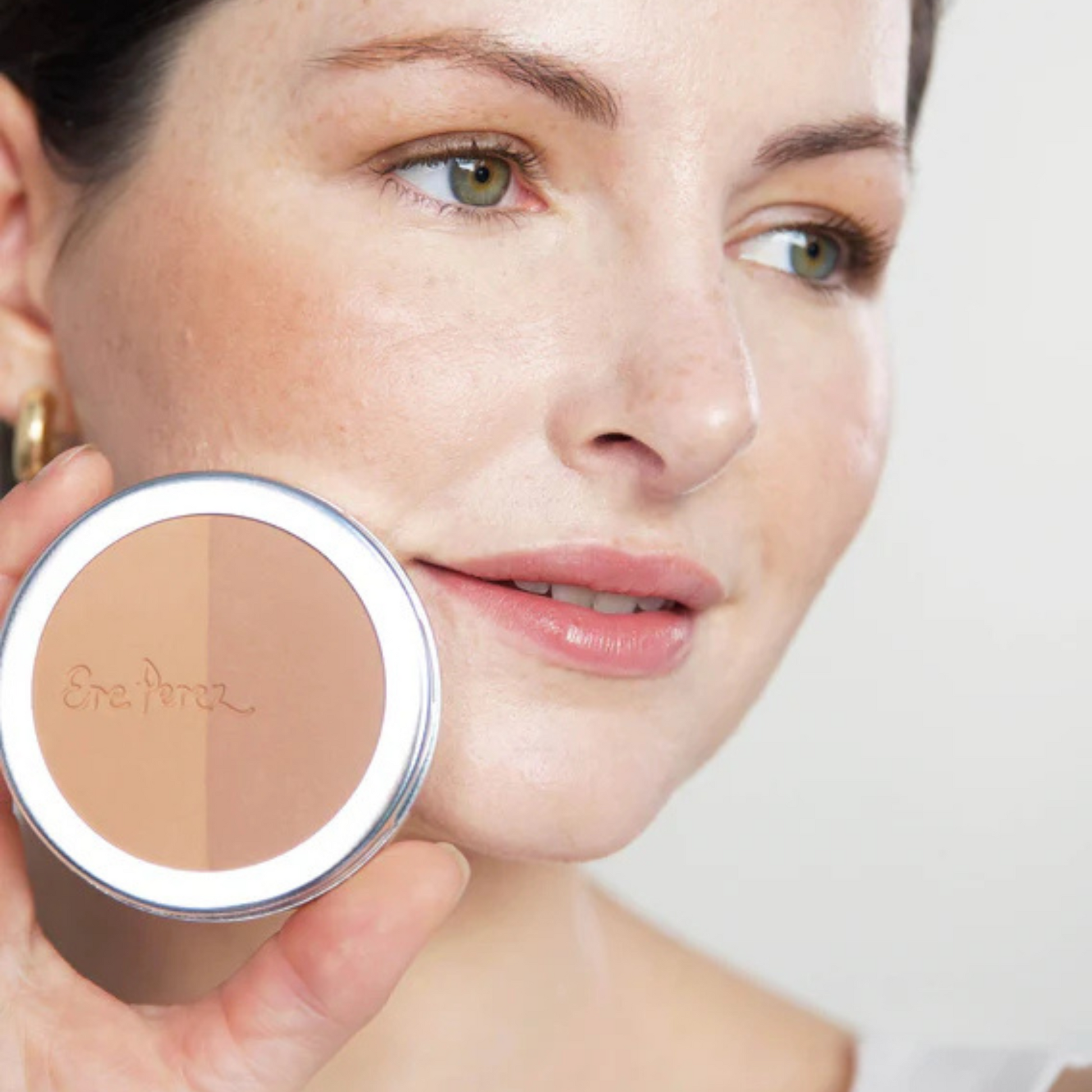 mujer sonriendo con tono roma en sus mejillas de blush y bronceador ere perez  sosteniendo envase