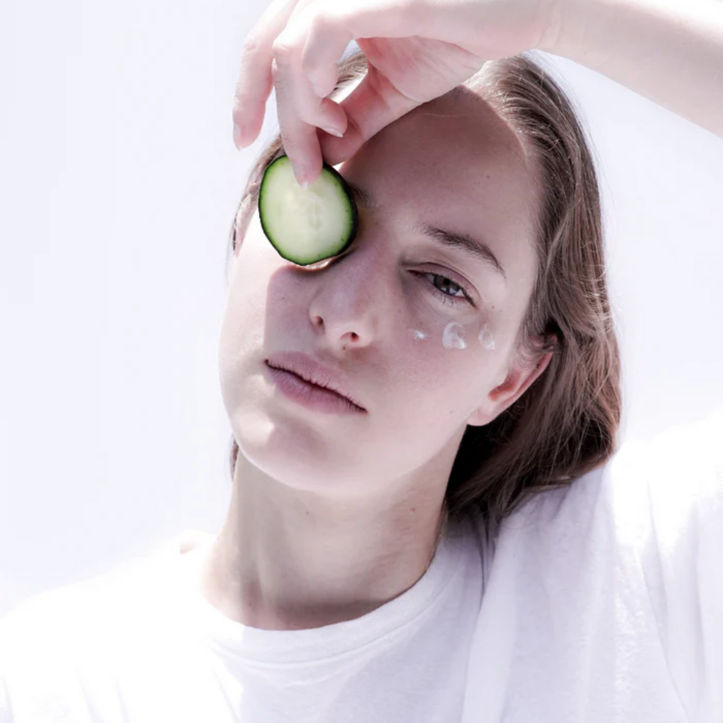 retrato de modelo con una pequeña cantidad de suero de ojos antiedad aplicada en las ojeras mientras sostiene una rodaja de pepino